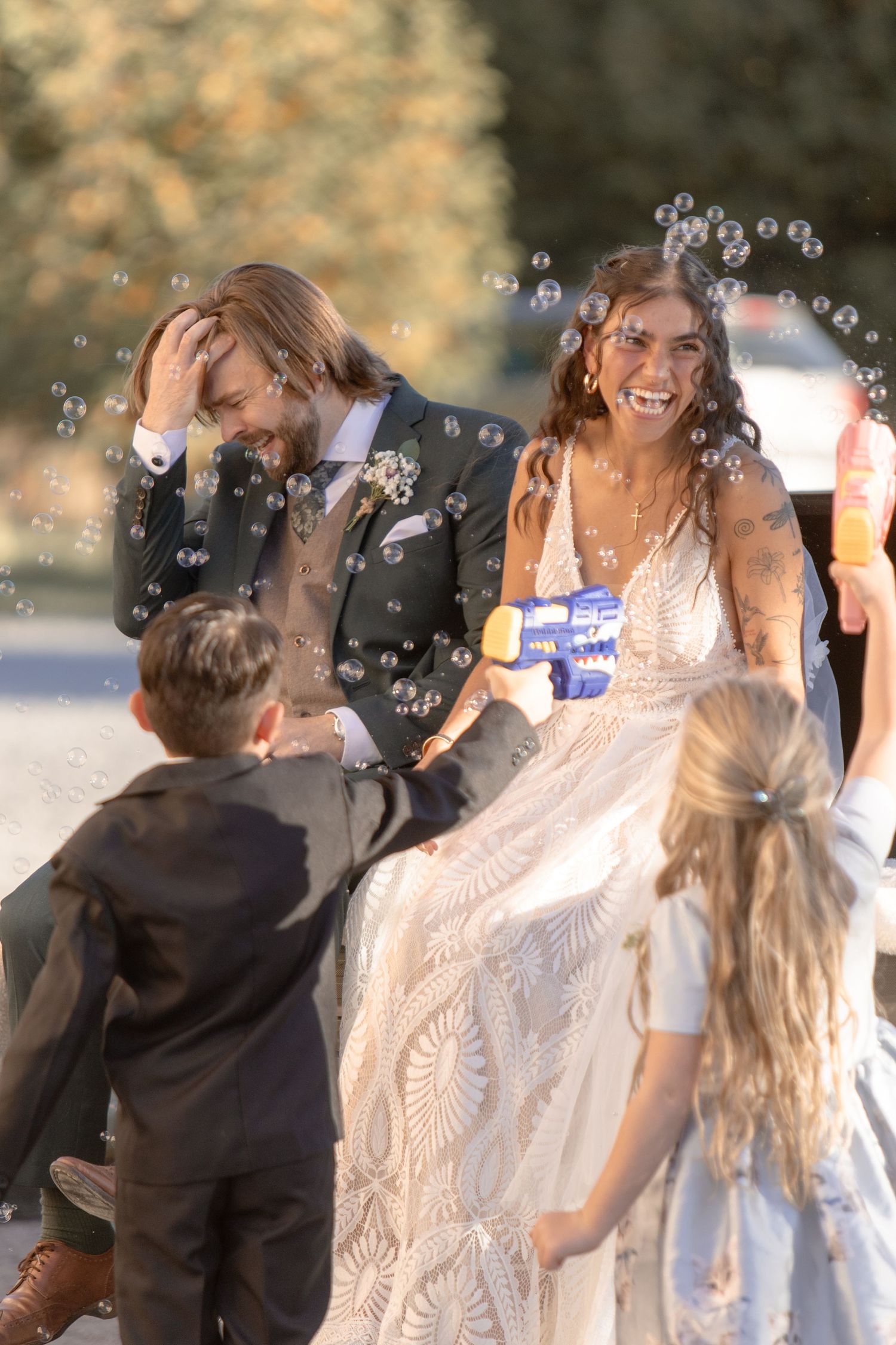 Wedding celebration with bubbles floating in the air as children play around a couple in a lace white dress outdoors.