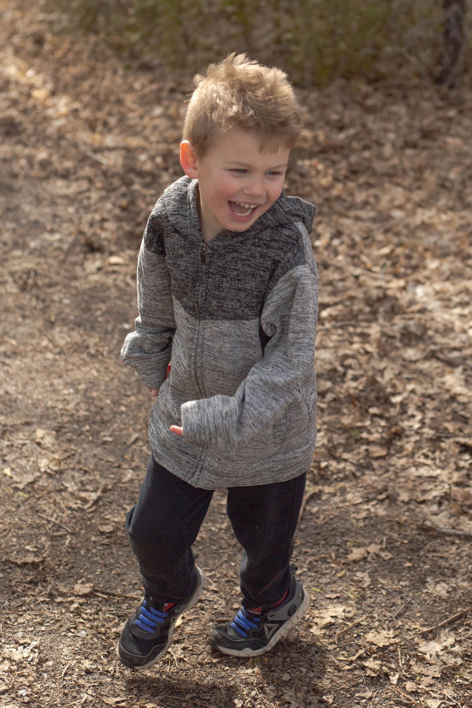 Child in grey hoodie plays energetically among fallen leaves on a forest trail wearing blue sneakers.