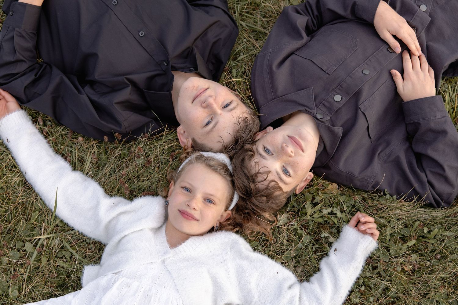 Three people lying on grass in a circle formation wearing dark jackets and white clothing.