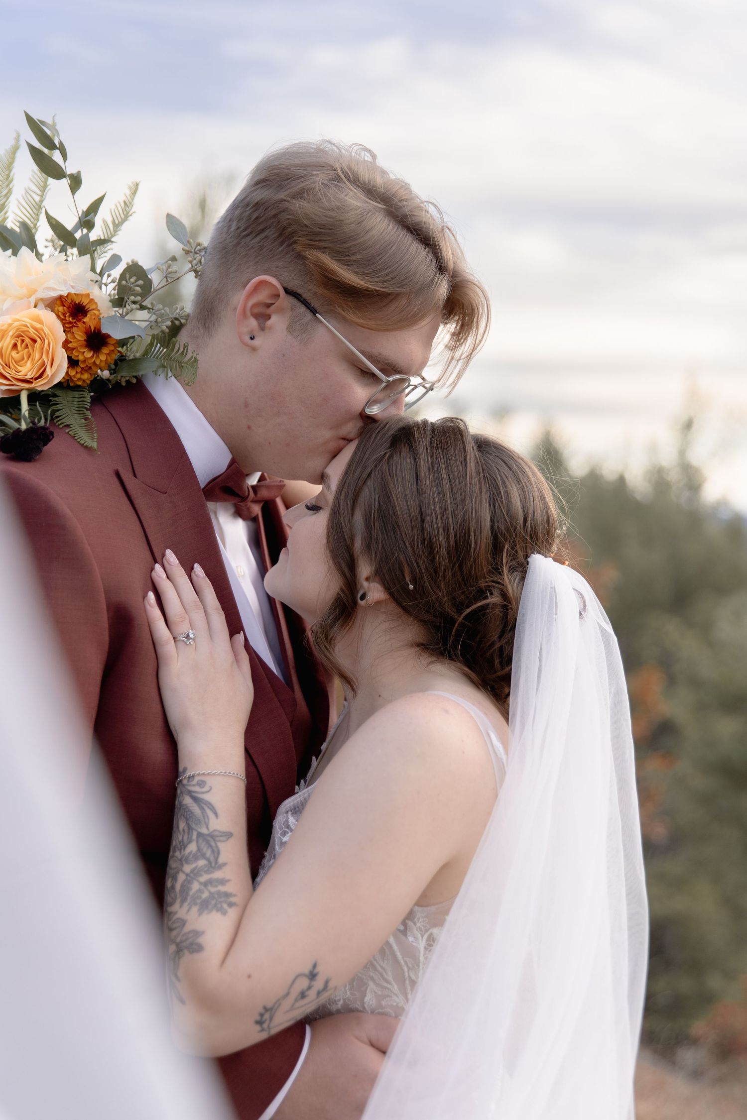 An intimate wedding moment captured as newlyweds share tender kisses against a soft cloudy sky backdrop.