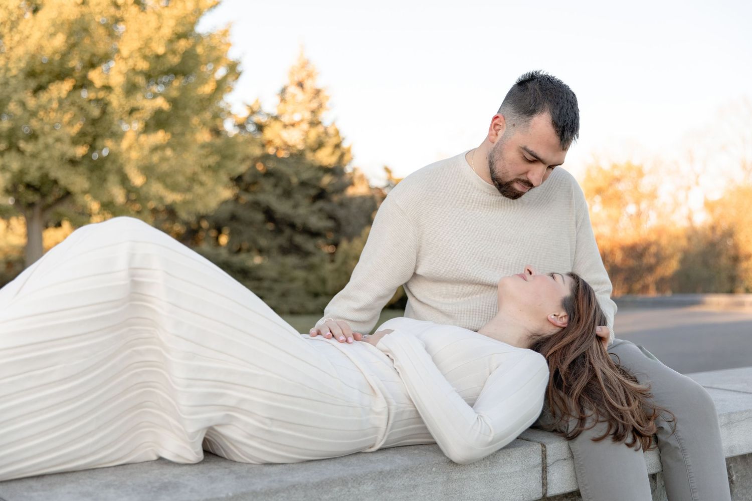A couple in cream colored sweaters embraces lovingly on a sun-drenched road during golden hour.