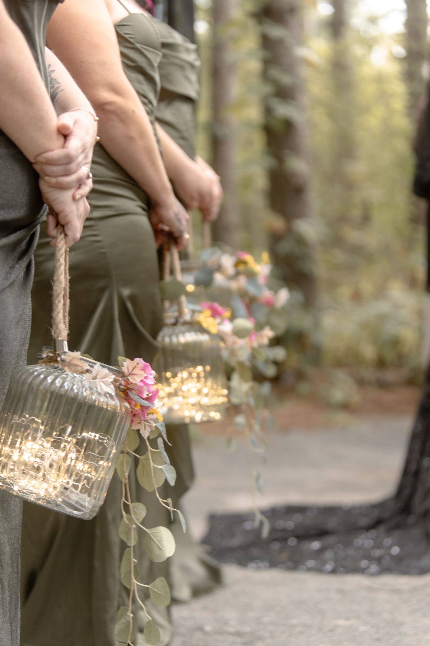 A person in olive green pants holds a glass jar with dried flowers and stems against a blurred forest background.