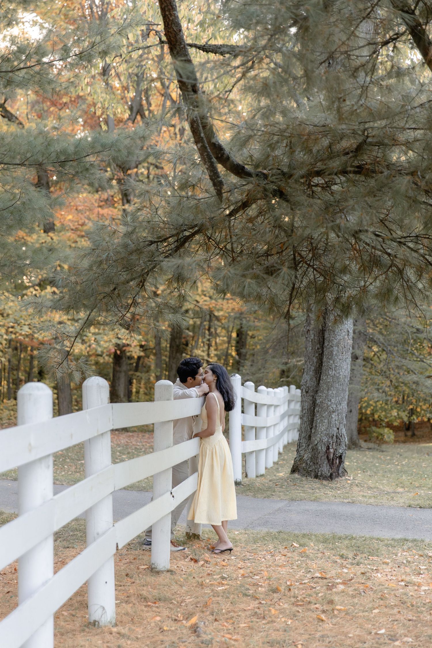A romantic moment captured by a white fence in an autumn forest with fallen leaves on the ground.