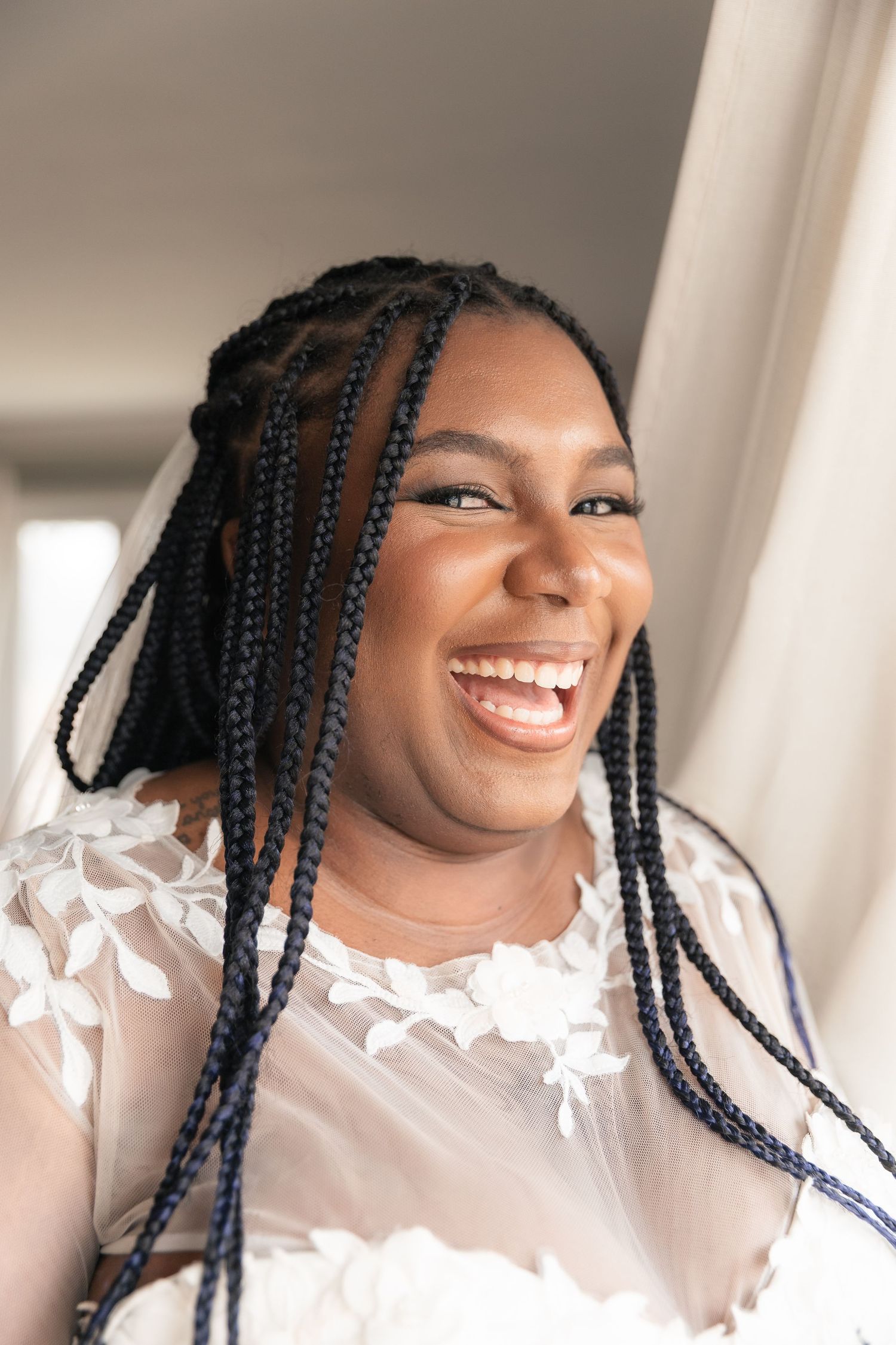 A joyful Black bride in a white floral gown with box braids and a veil, laughing radiantly near a window.