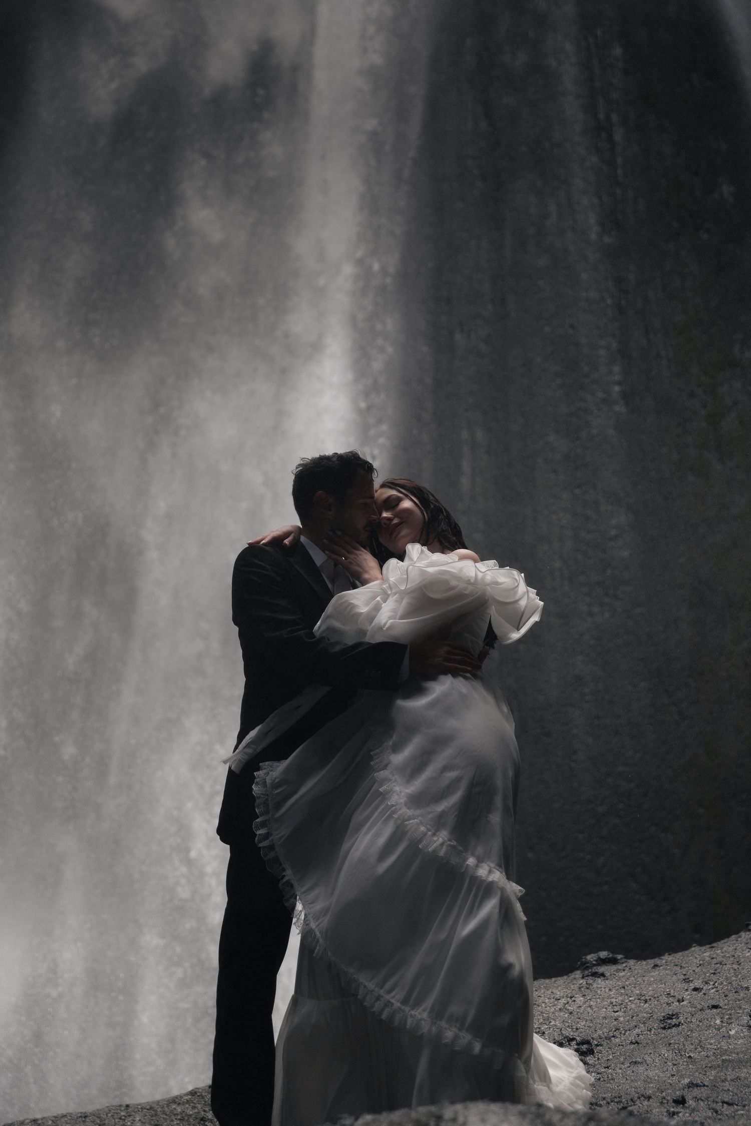 Wedding couple share a tender kiss and embrace before a stunning misty waterfall backdrop.