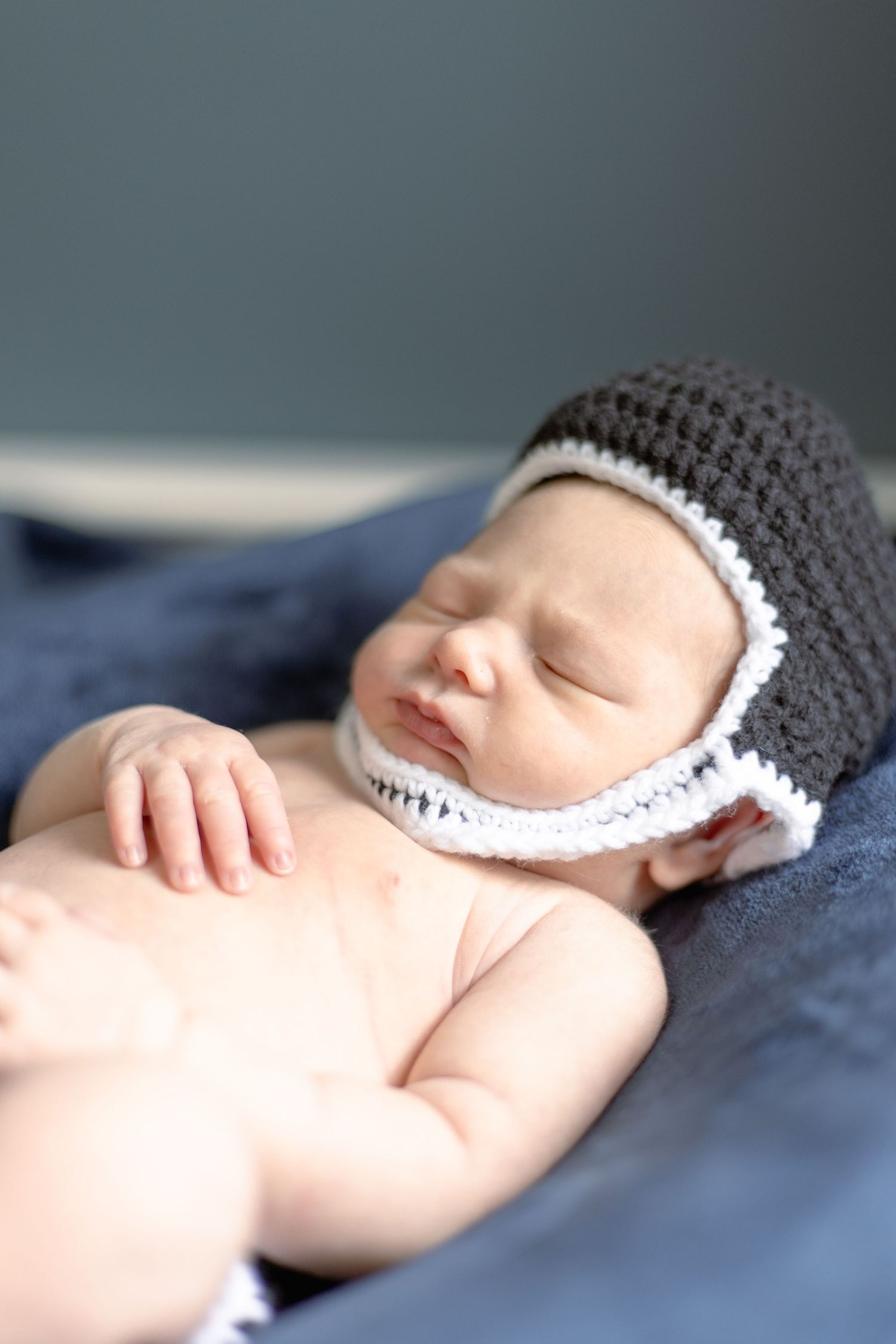 A sleeping newborn wearing a gray and white crocheted bonnet lies on dark blue fabric.
