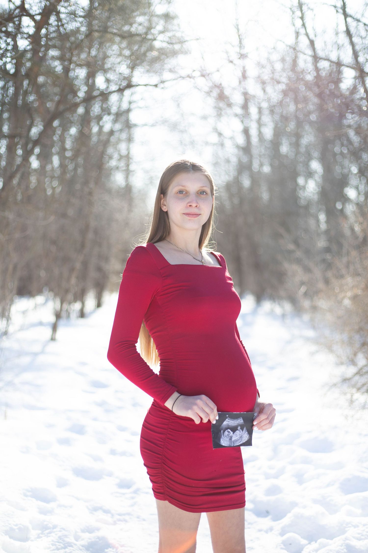 Winter maternity shoot featuring red dress and ultrasound photo in snowy setting.
