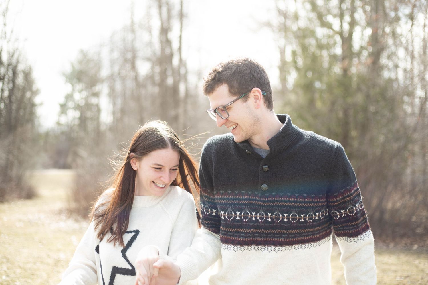 A couple in cozy winter sweaters laugh together while walking outdoors in the soft sunlight.