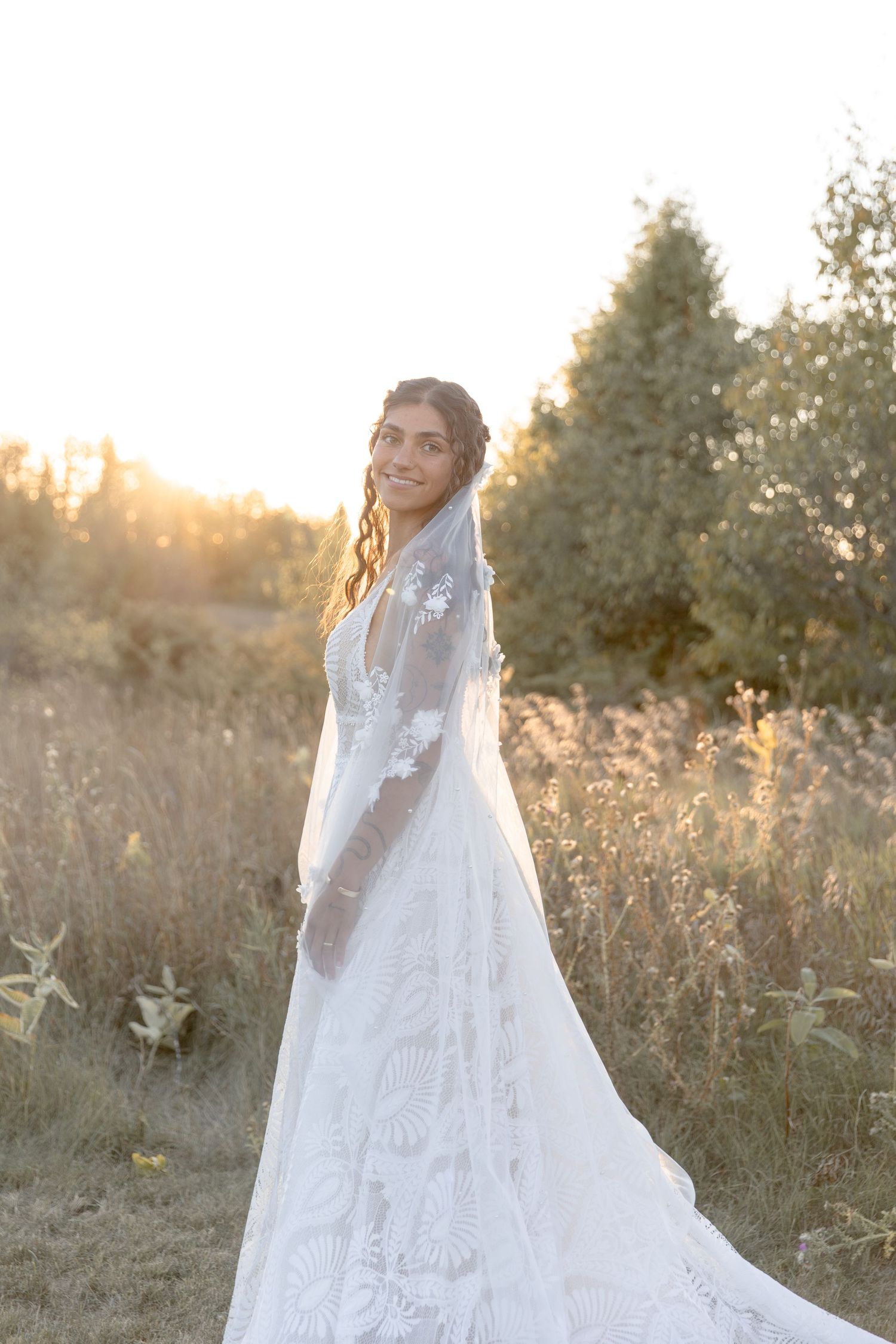 A bride in a flowing white lace wedding dress poses in a sunlit meadow during golden hour.