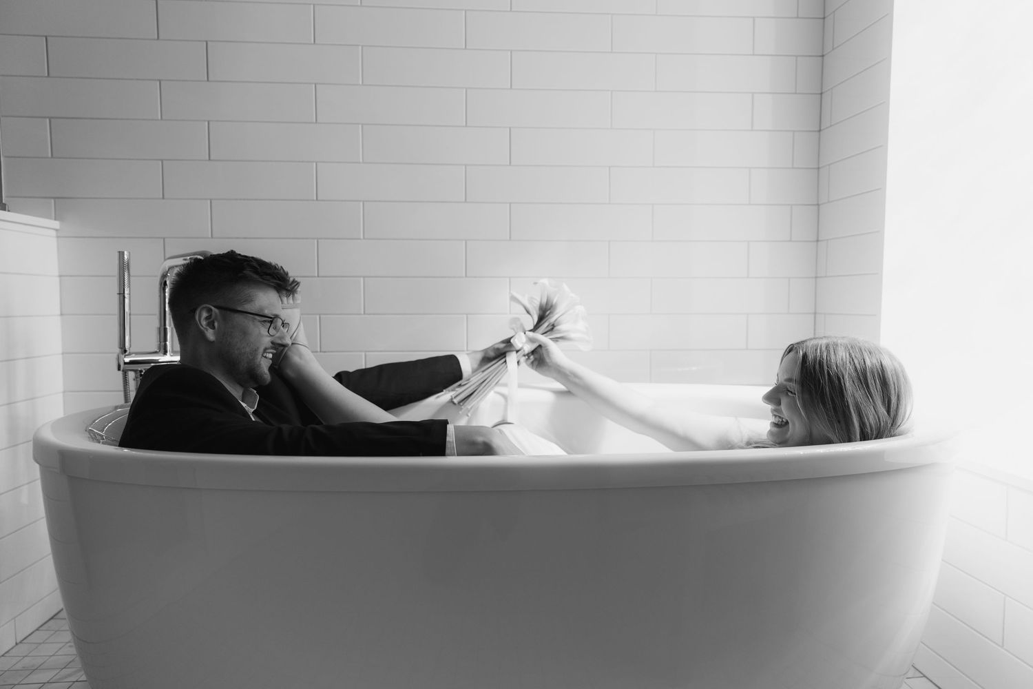 A couple shares a playful moment together in a white freestanding bathtub.