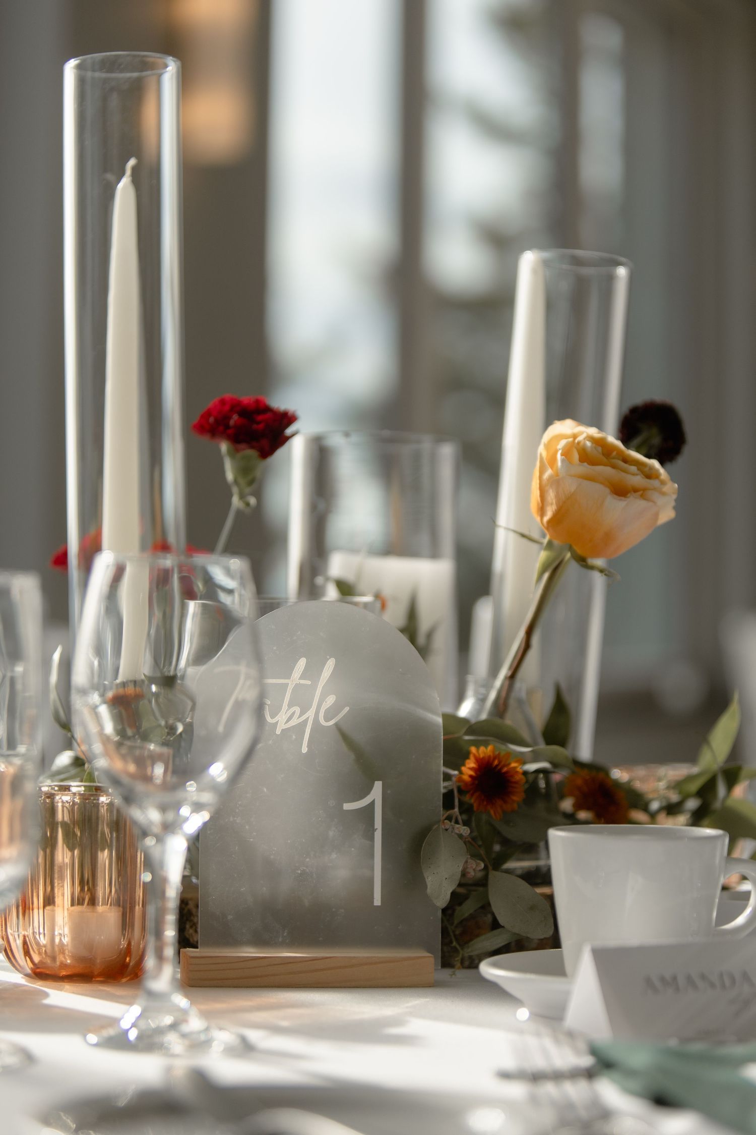 Elegant wedding table settings with white candles, delicate glassware, and scattered floral accents in soft natural light.