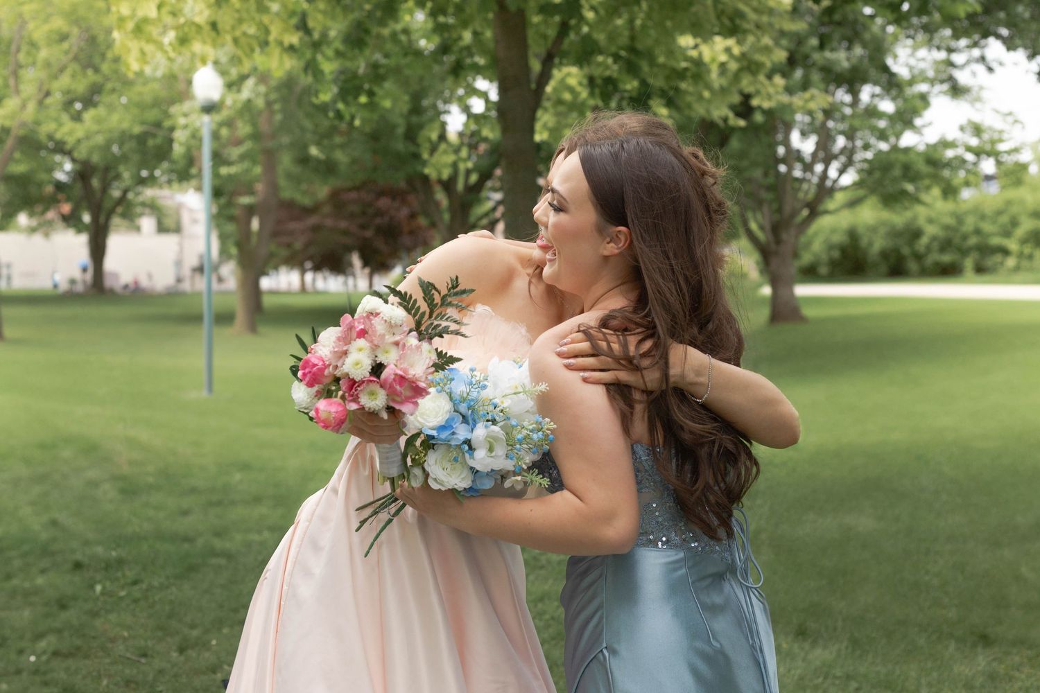 Two women in formal dresses embrace while holding a pink and blue floral bouquet in a sunny park setting.