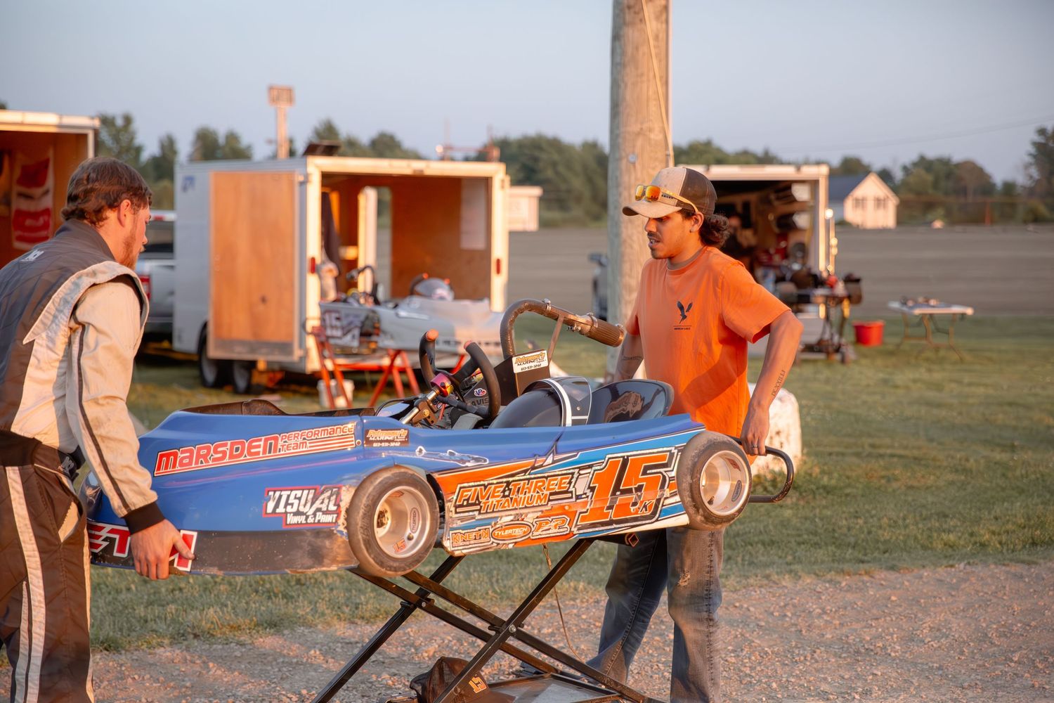 Two people preparing a miniature racing car on a workbench at an outdoor track during sunset.