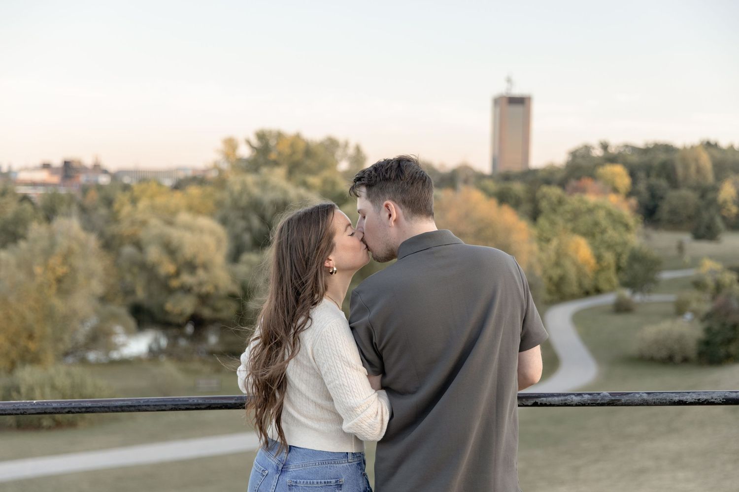 A couple shares a romantic moment at sunset with a cityscape and autumn trees in the background.