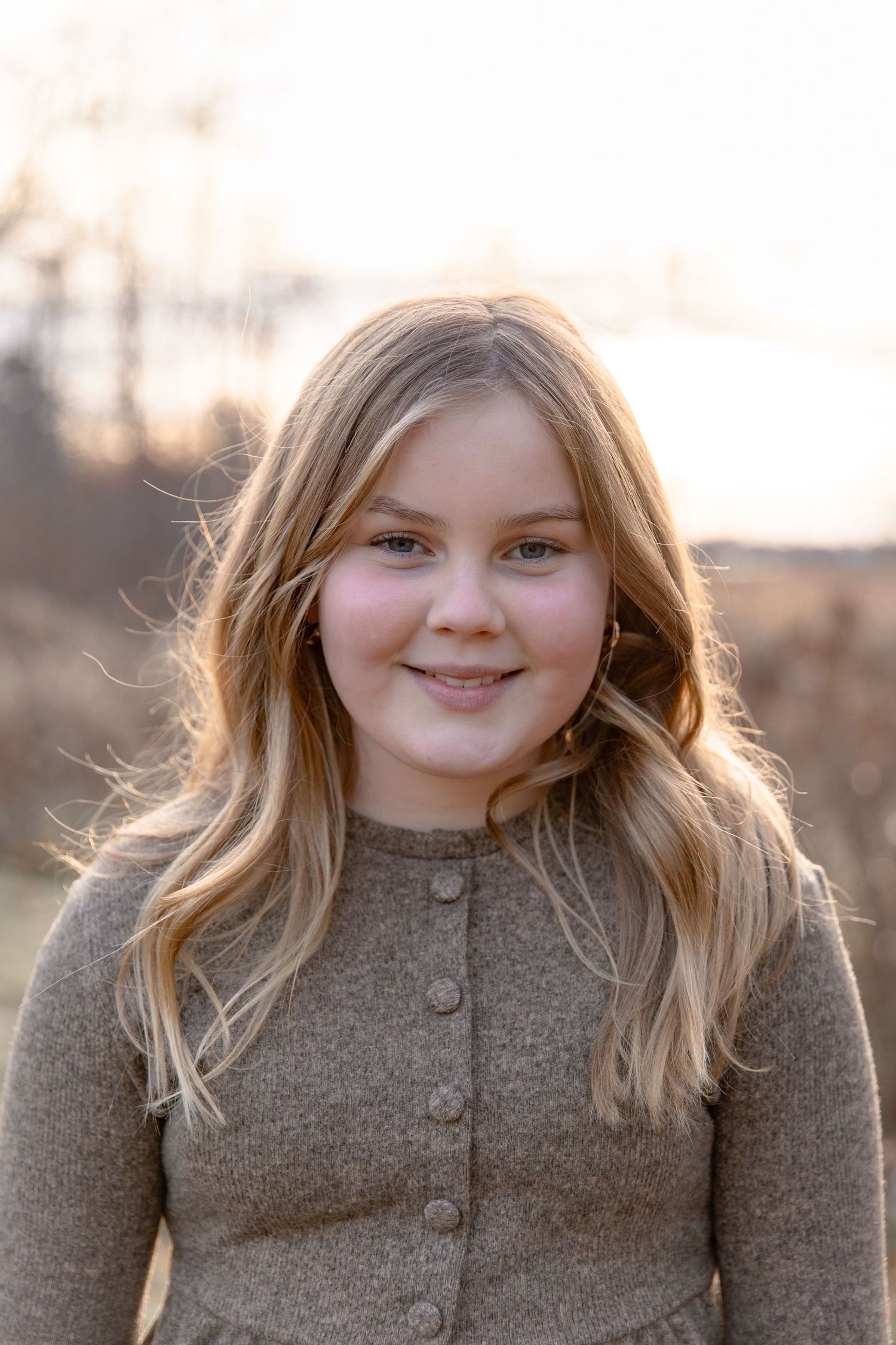 A young person wearing a brown knit sweater smiles warmly while standing outdoors during golden hour.