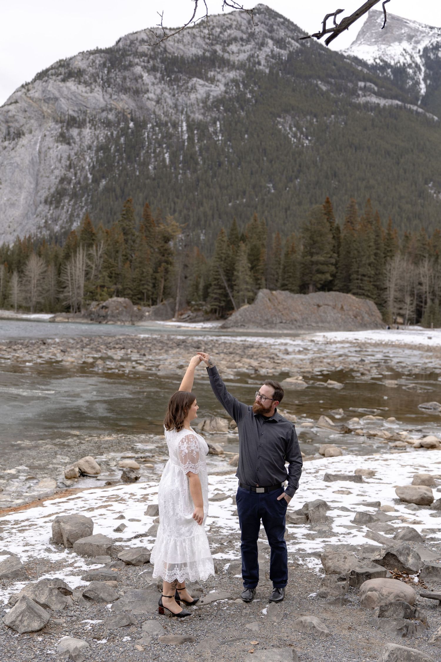 Two people dance together on rocky riverbank with majestic snow-capped mountain and evergreen forest in background.