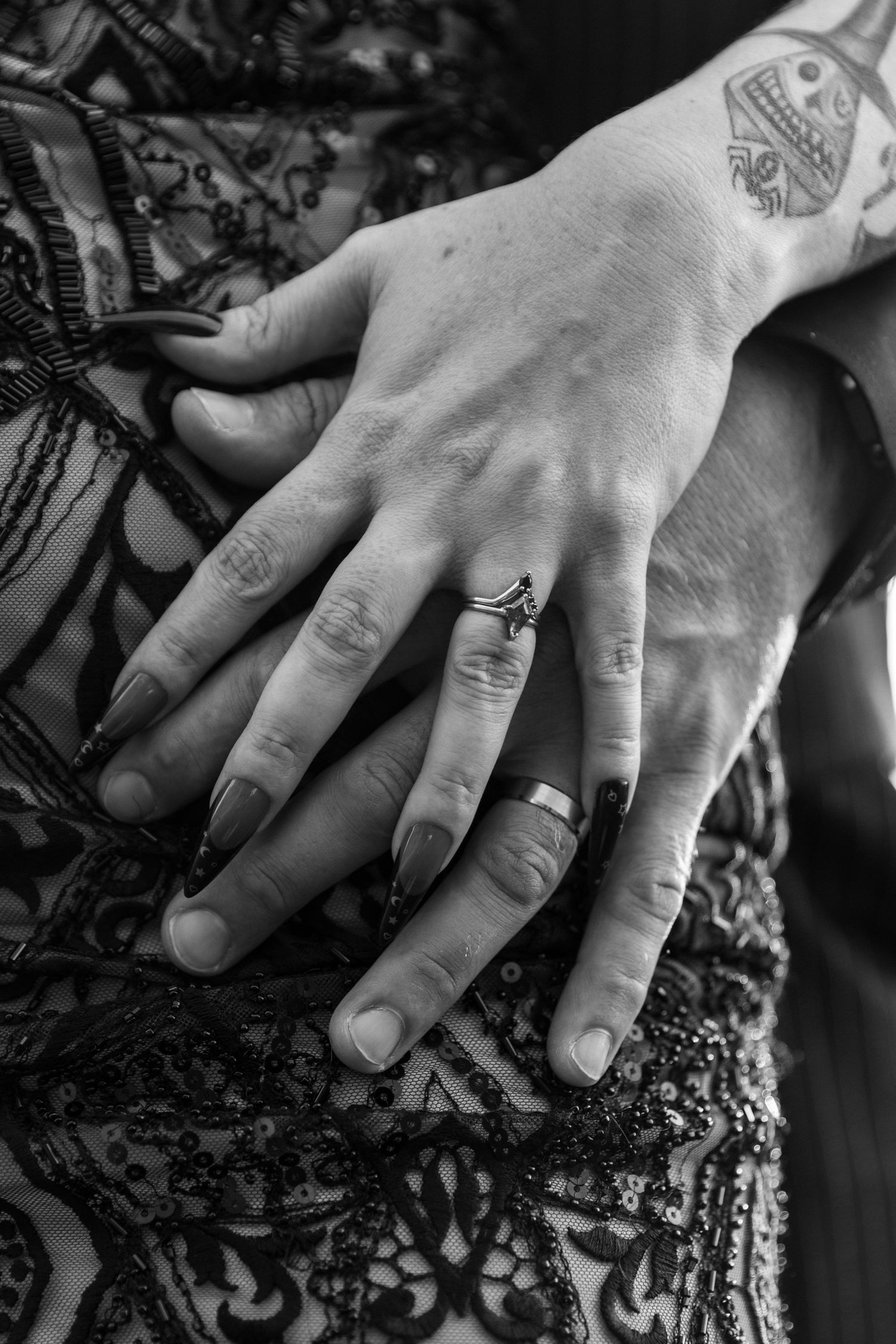 Black and white close-up of hands displaying wedding rings against a lace pattern background.