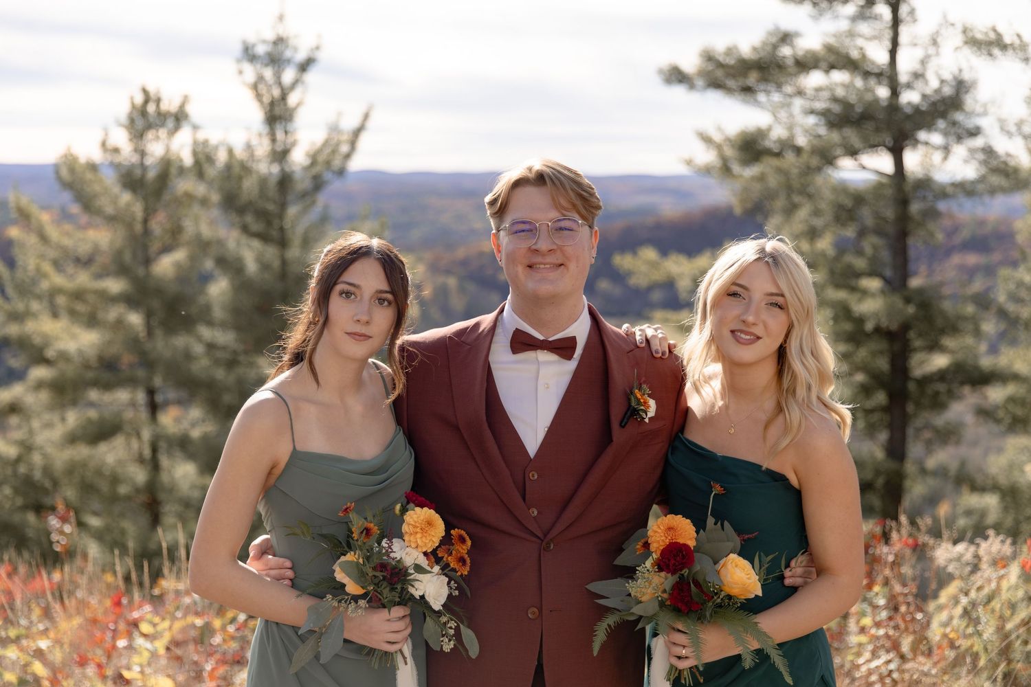 Wedding party poses outdoors in burgundy and sage green attire against a scenic mountain backdrop on an autumn day.
