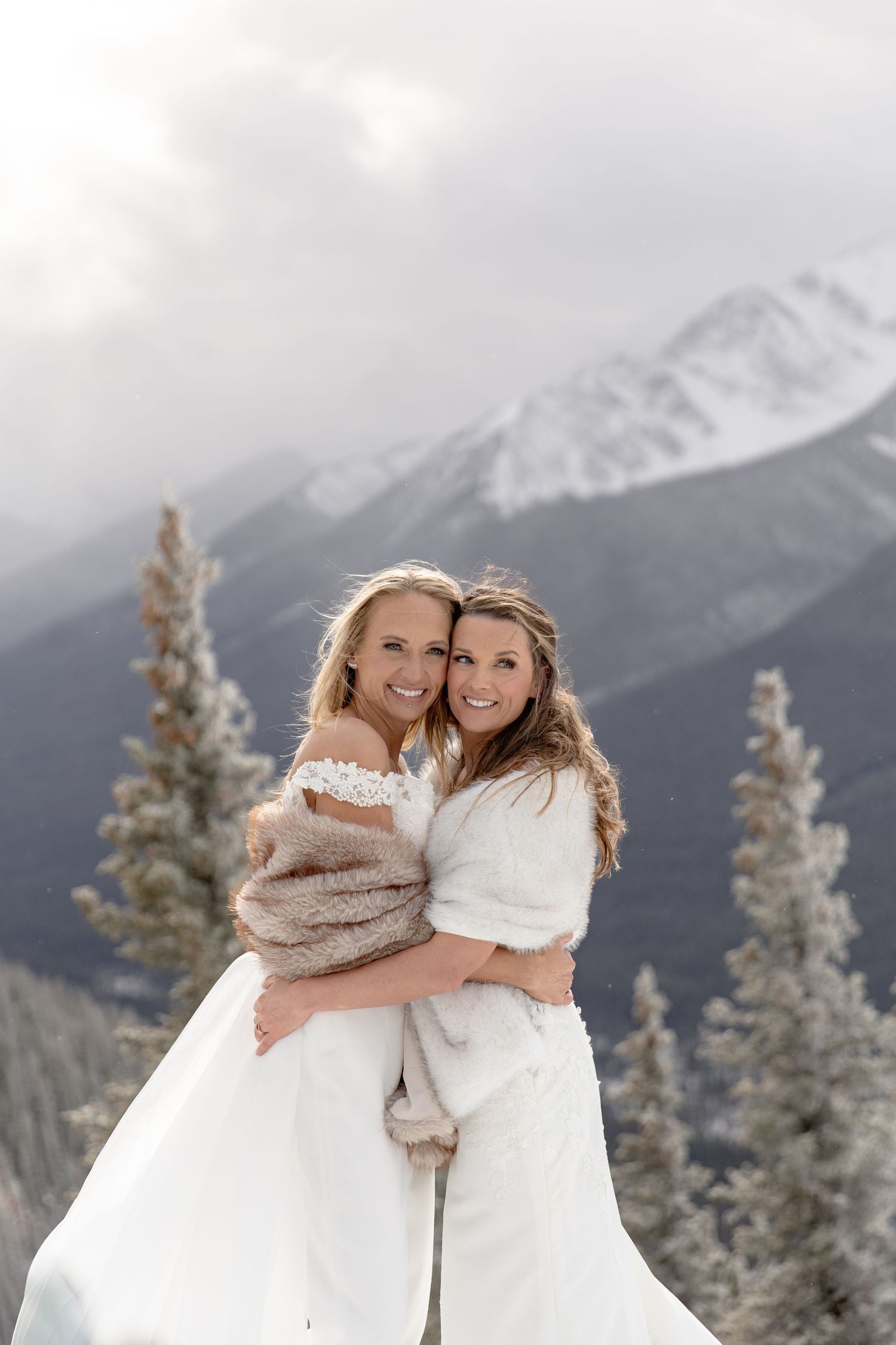 Two people in white wedding attire share an intimate moment with snow-covered mountains and pine trees behind them.