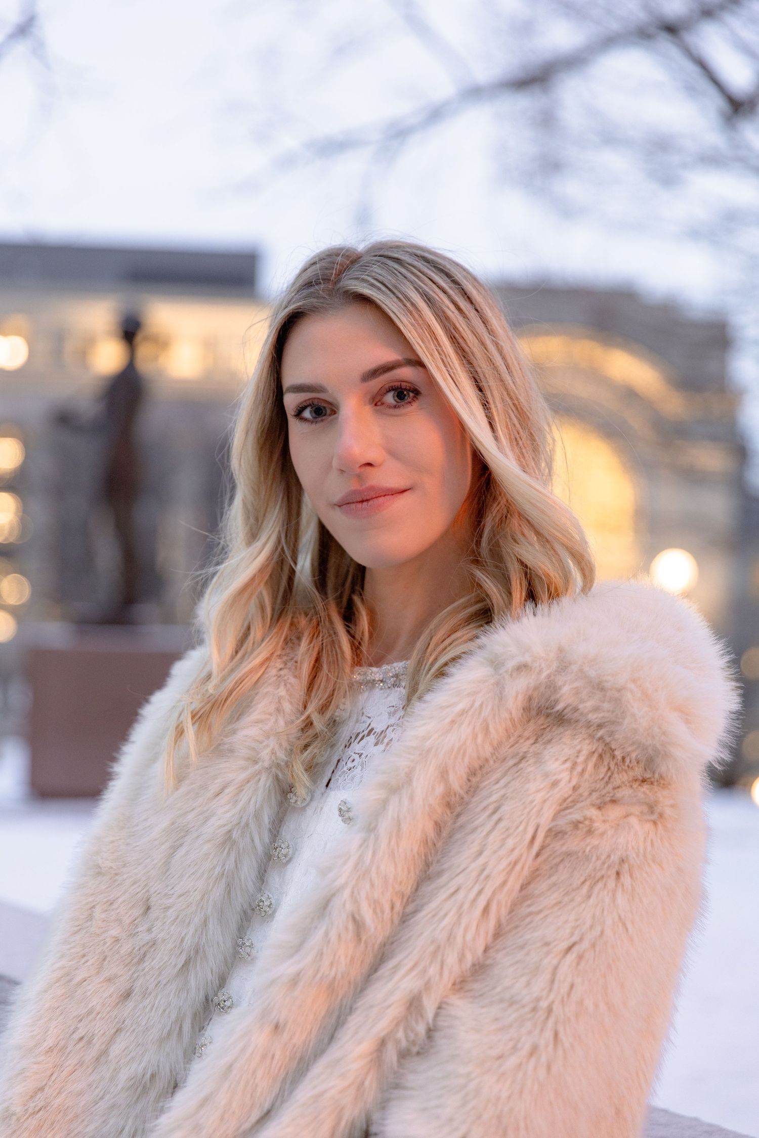 Woman in a plush cream-colored faux fur coat poses outdoors in winter with soft lighting and bokeh effects.