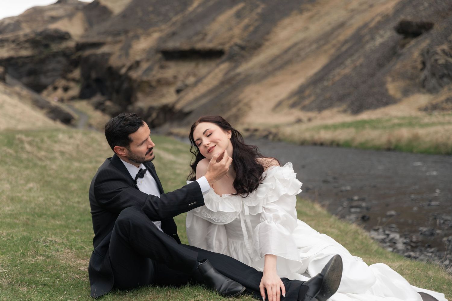 Couple in wedding attire sitting on grass by a stream with dramatic mountain backdrop in Iceland.