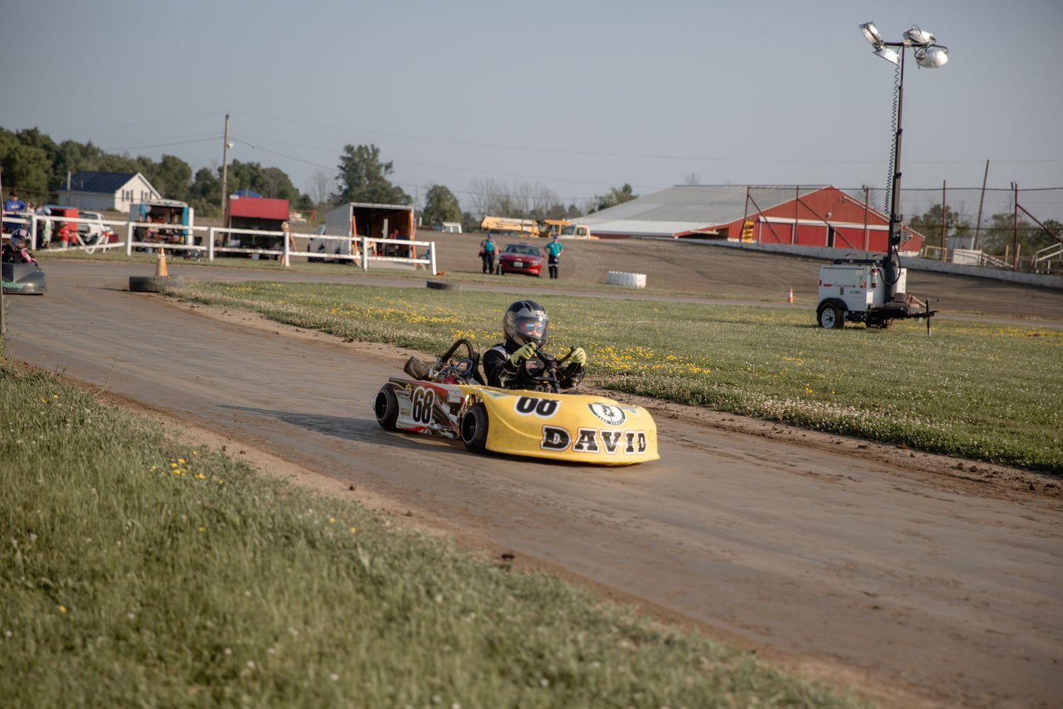 Yellow go-kart racing on an outdoor track at a motorsports facility with red buildings in the background.