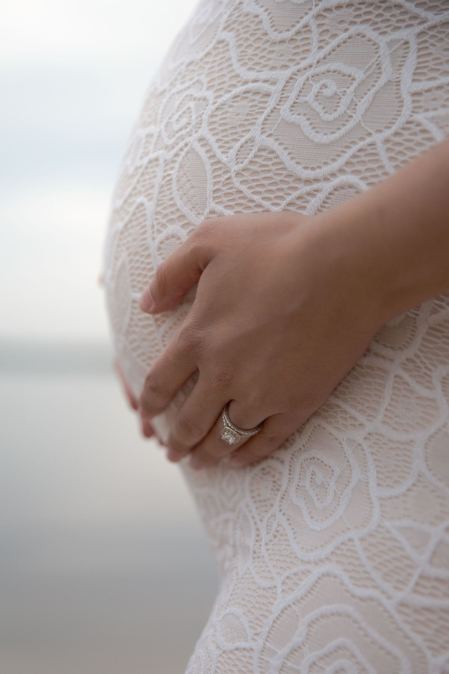 Close-up of a hand with engagement ring resting on white lace dress fabric against a soft blurred background.