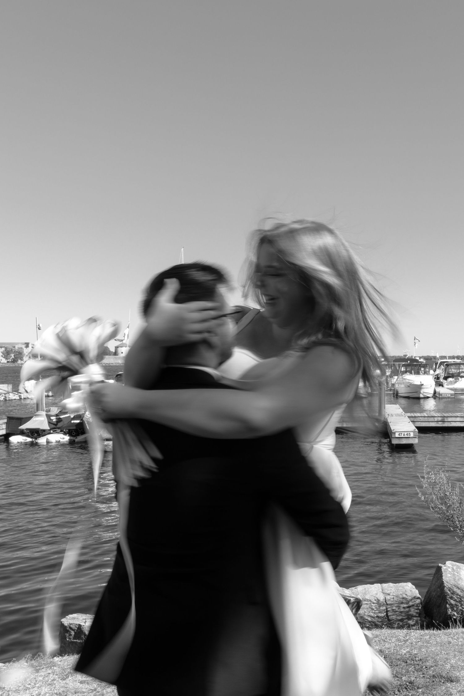 A couple embraces and spins joyfully by a marina on a sunny day, with boats in the harbor visible in background.