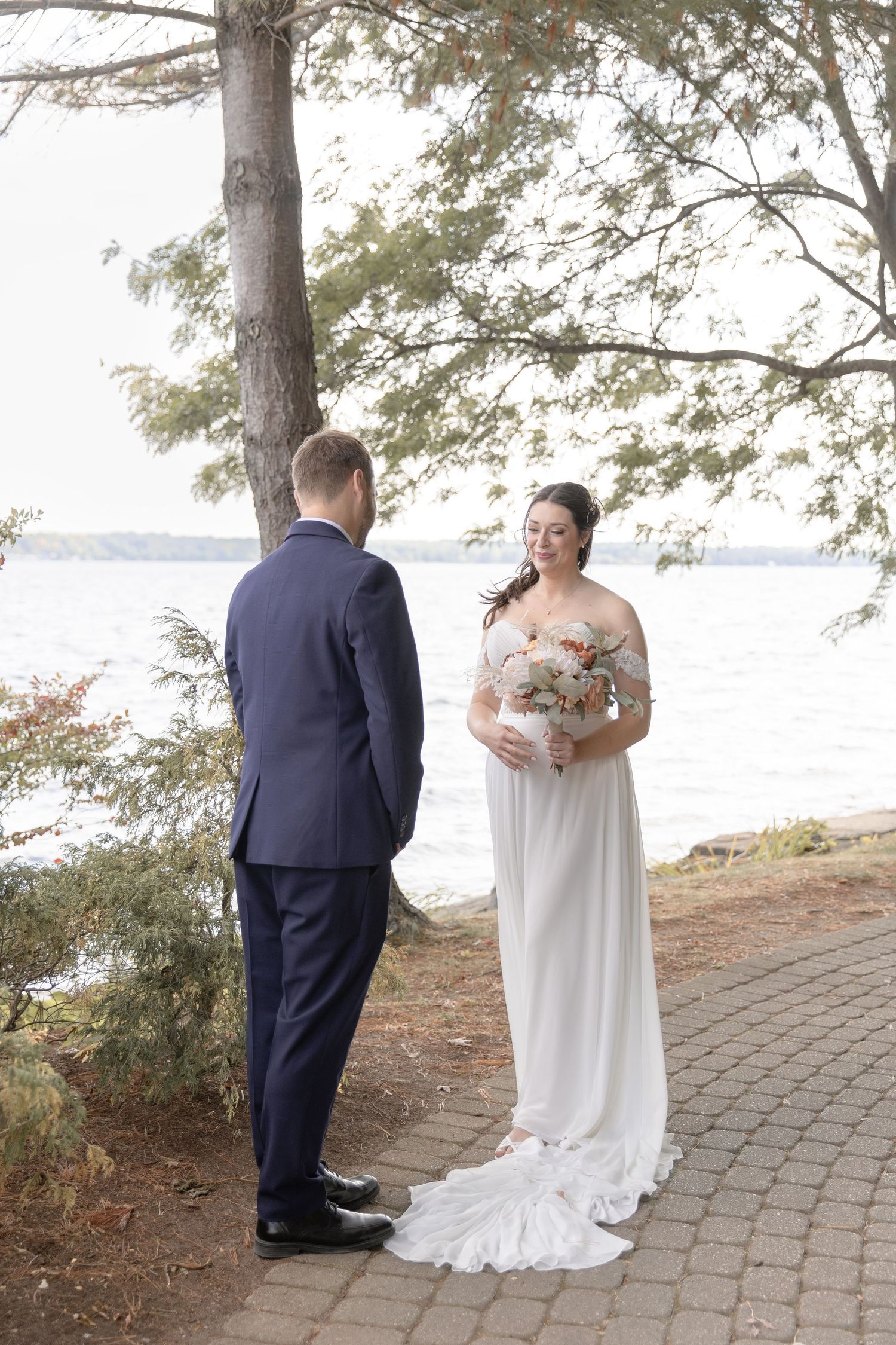 A couple in wedding attire shares a romantic first look moment on a brick path by a lakeside under tall pine trees.