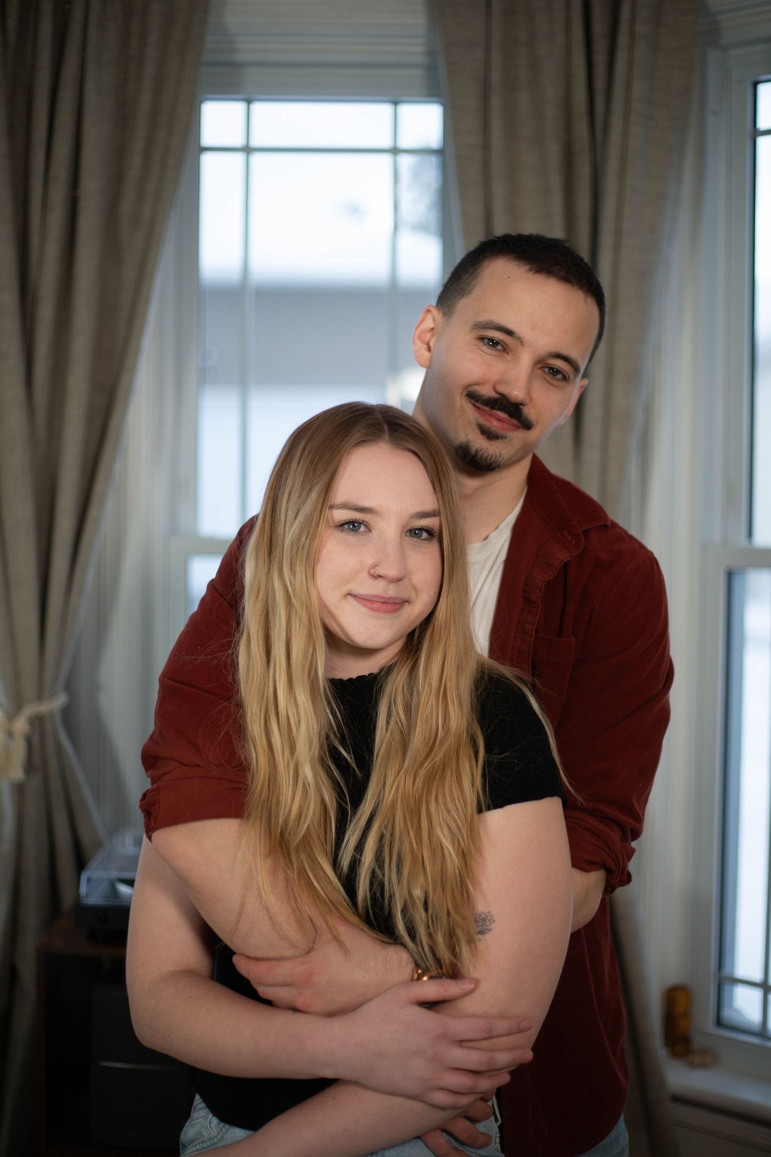 Tender embrace of couple wearing red and black against window light.
