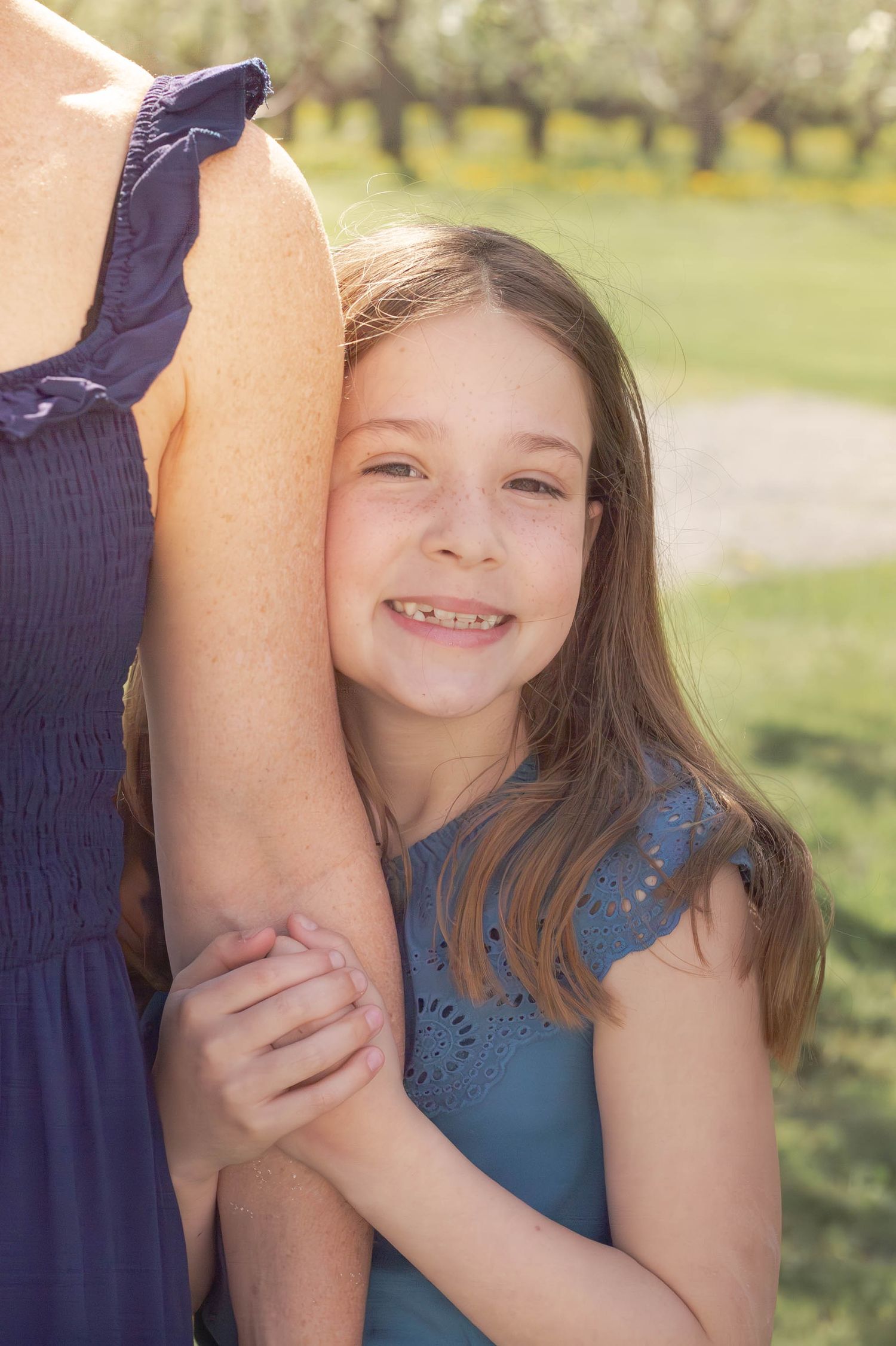 Young girl in a blue dress hugging an adult's arm while standing in a sunny outdoor setting.
