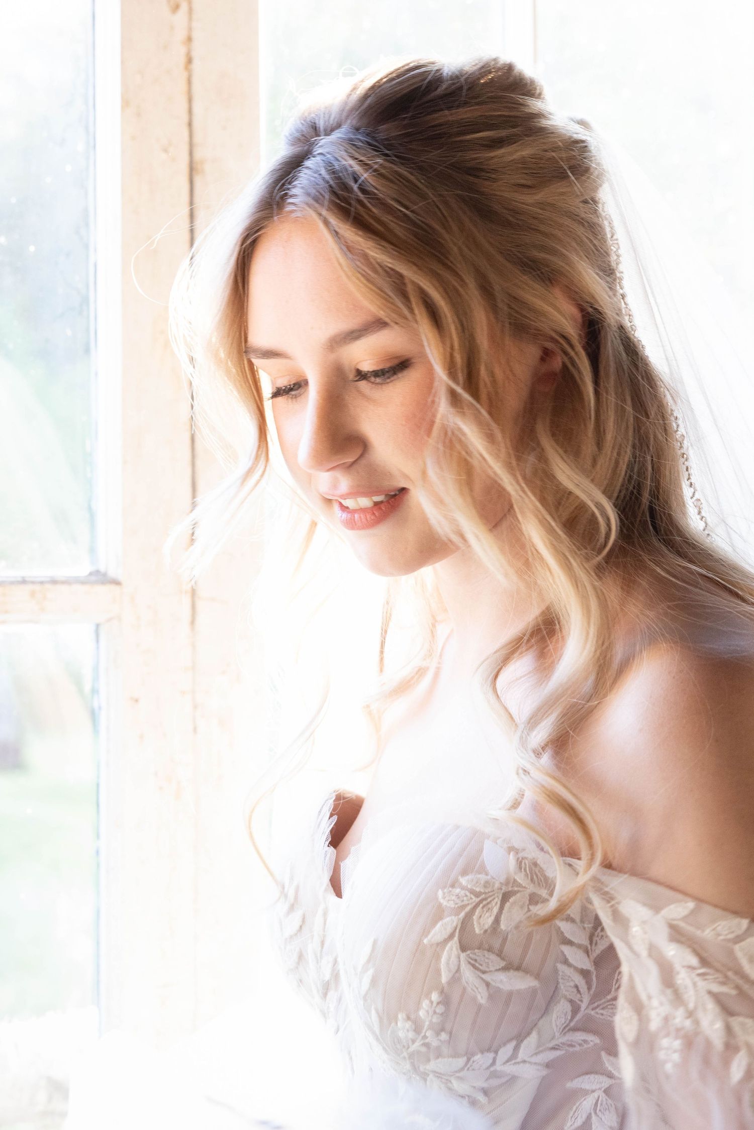 A gentle smile graces a woman in an off-shoulder white lace dress by a sunlit window, with soft blonde wavy hair.