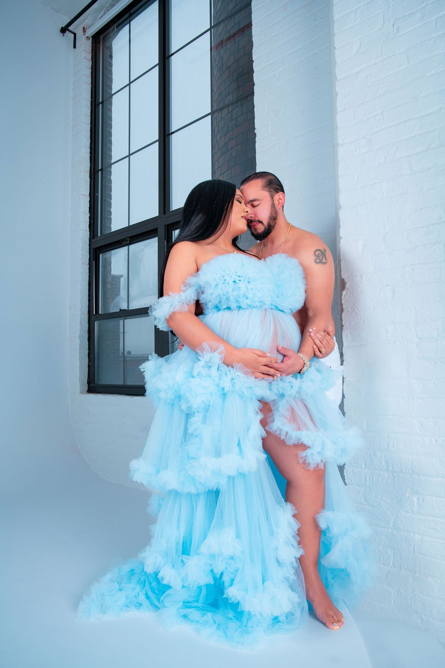 Couple embraces in light blue tulle gown during romantic maternity photoshoot.