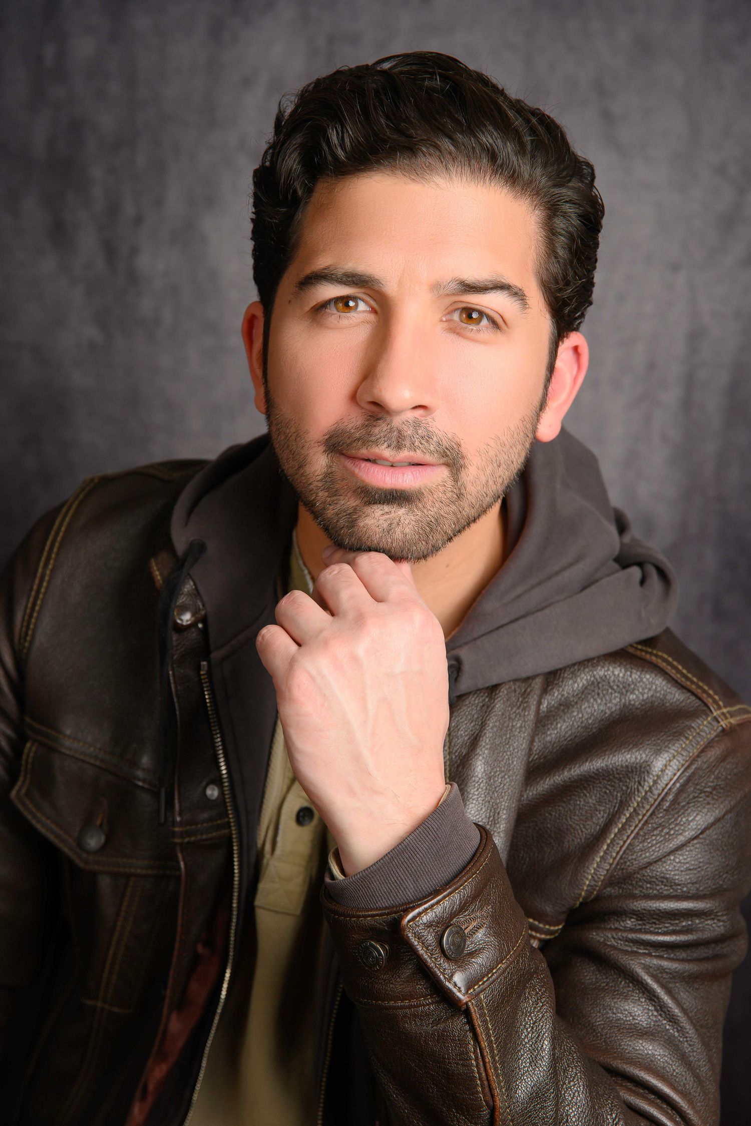 Professional headshot portrait of someone in a brown leather jacket and gray hoodie with hand on chin against dark background.