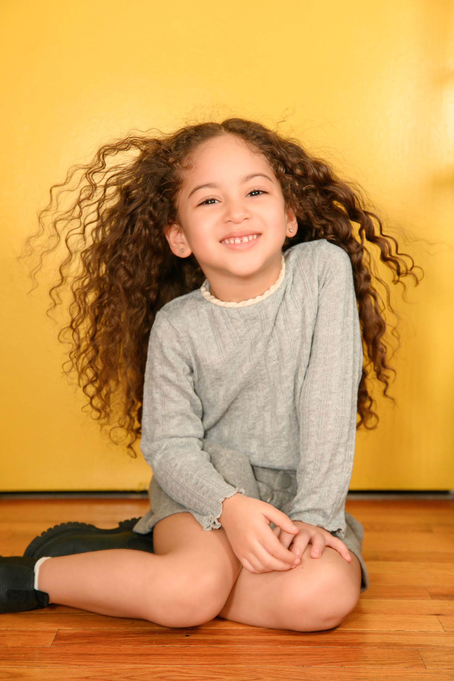 Child with curly hair wearing grey sweater sits on wooden floor against yellow background.