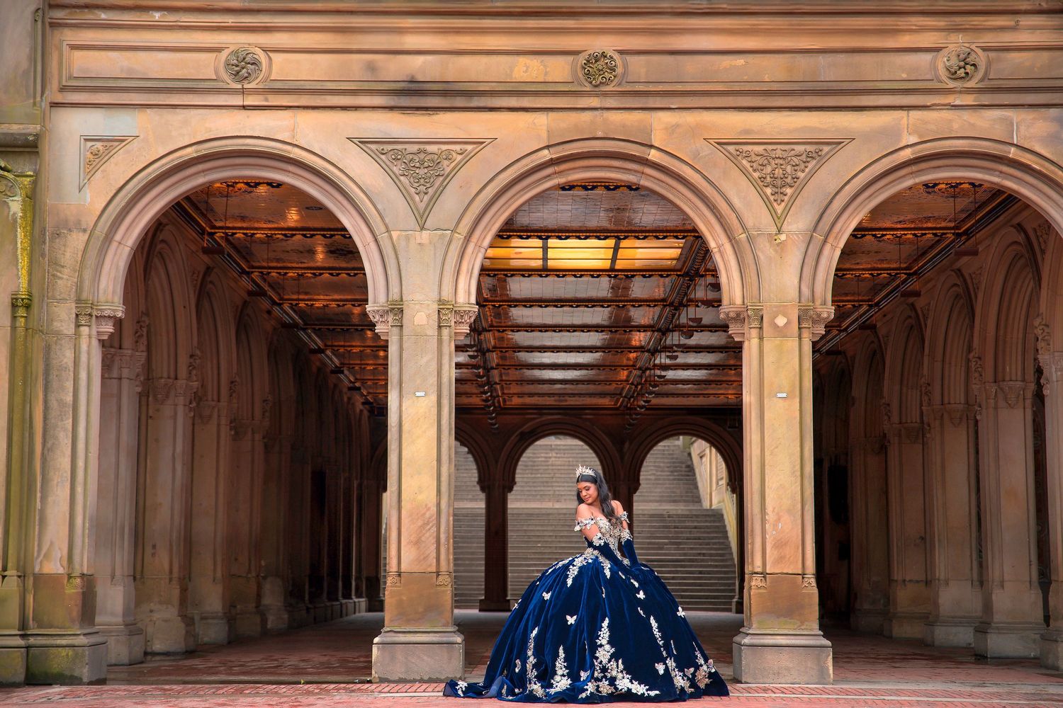A person in an elegant blue ball gown poses under ornate stone archways at Bethesda Terrace in Central Park.