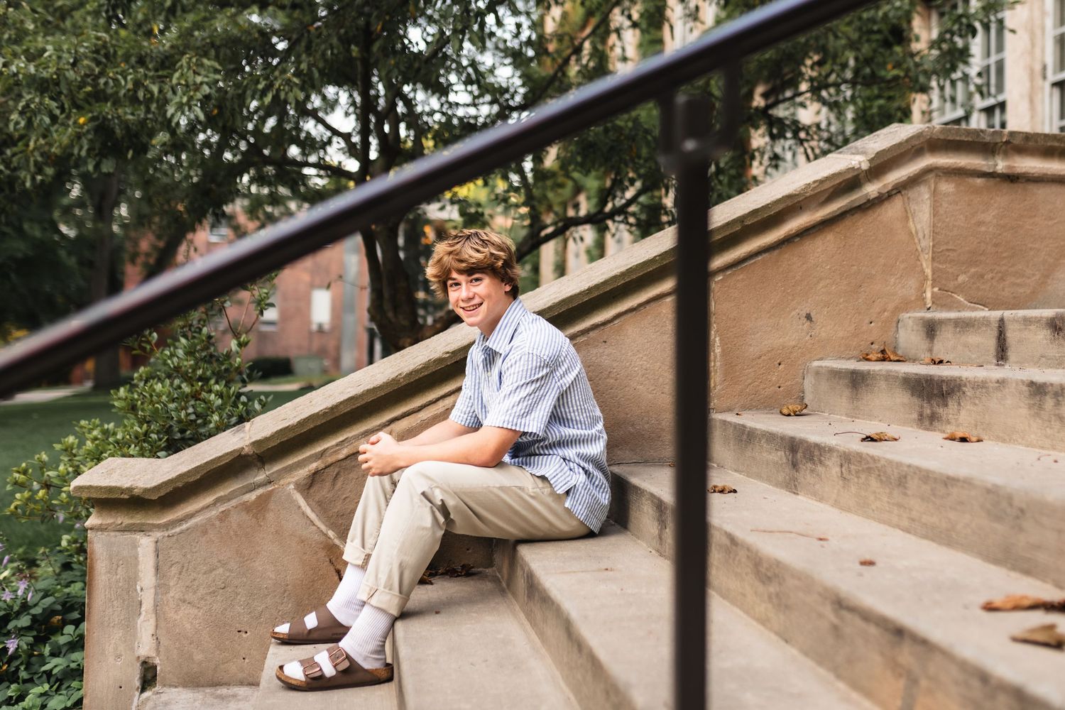 Senior portrait of a boy looking through the handrail while sitting on the steps at Aquinas College in Grand Rapids, MI