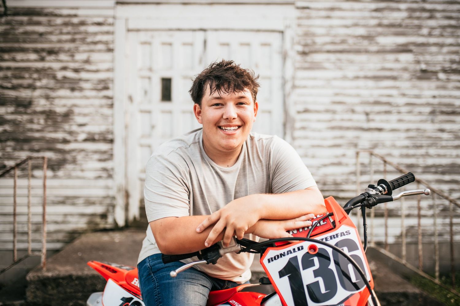 Senior portrait of a boy smiling while posing on his dirt bike, with an old rural church as a backdrop.
