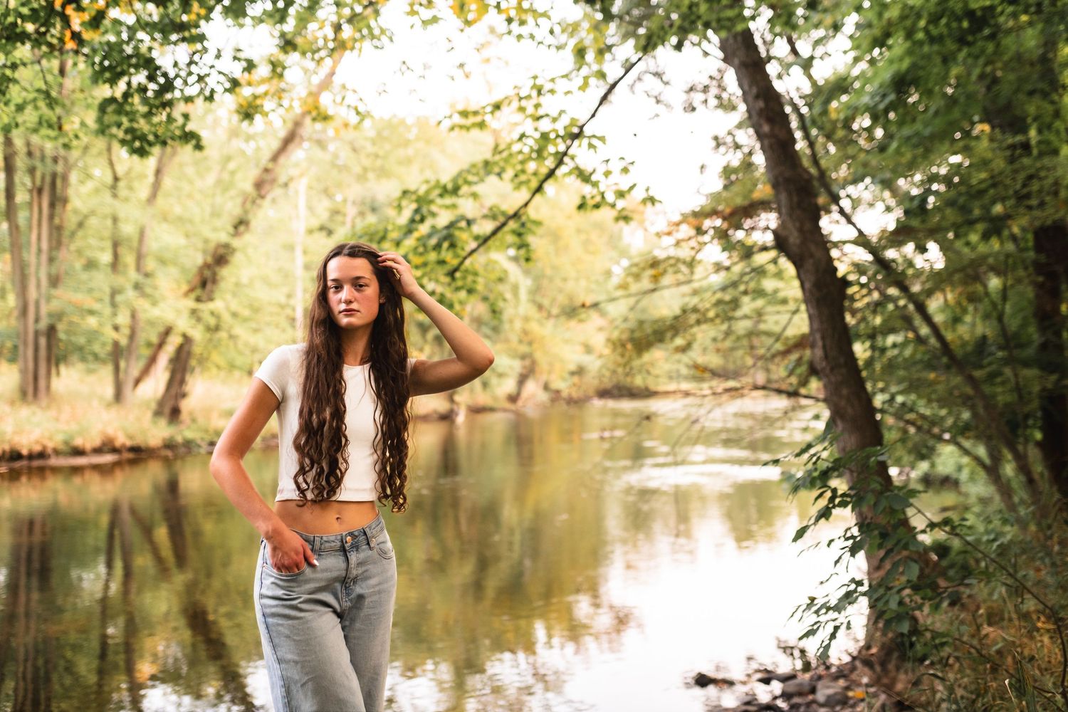 Senior portrait of a girl posed with her fingers through her hair with the Grand River in Portland, MI in the background.
