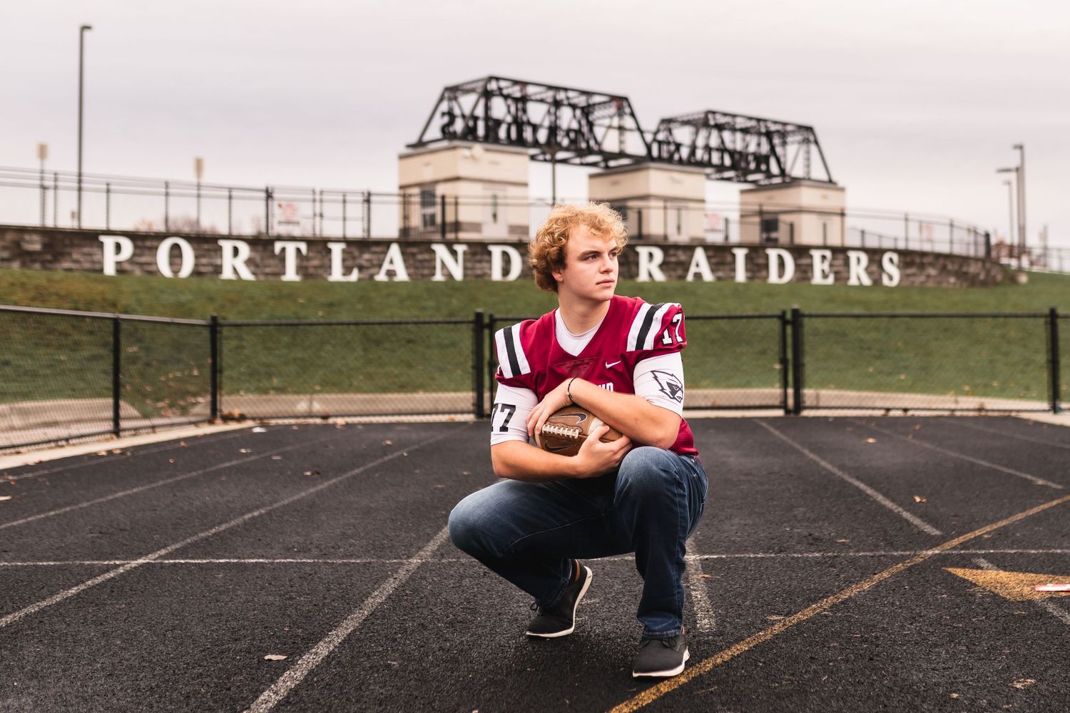A senior football player posing for his senior portrait while holding a football, with the stadium entrance in the background.