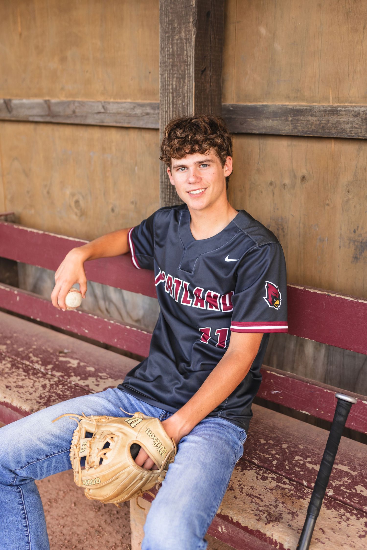 Portrait of a senior boy with a baseball and baseball mitt, posing in a dugout with his baseball jersey on.