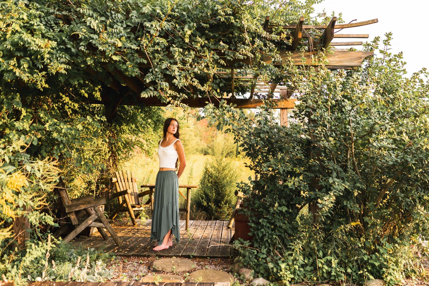 A senior girl looking off into the distance, standing in the shade of  an overgrown pergola
