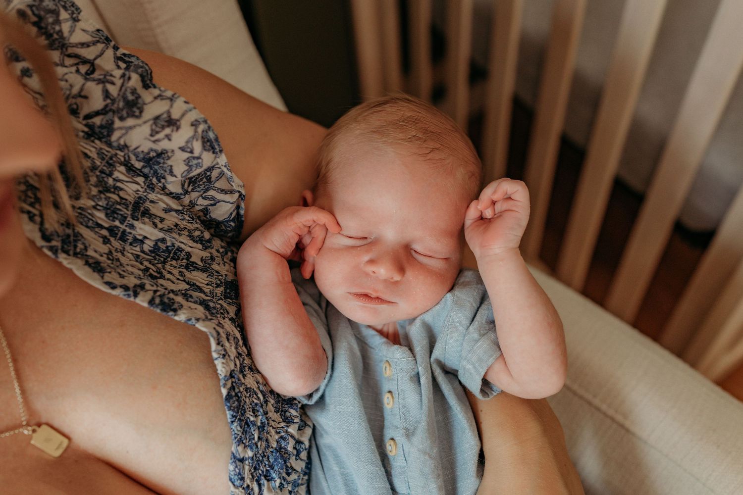 A sleeping infant wearing light blue clothing stretches on floral bedding in a white crib.