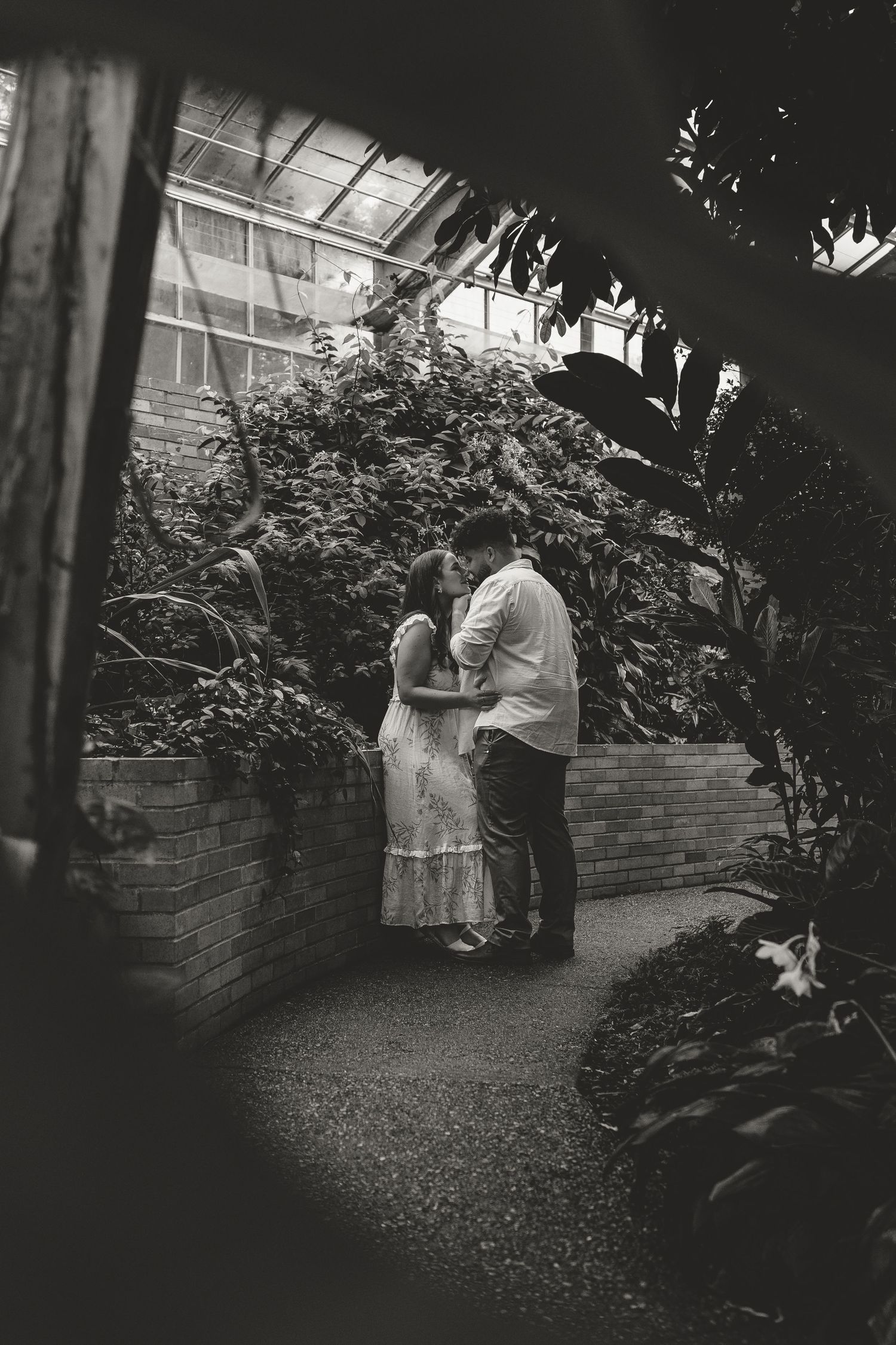 Two people embrace in a romantic moment beside a brick wall and garden shrubs in a black and white photograph.