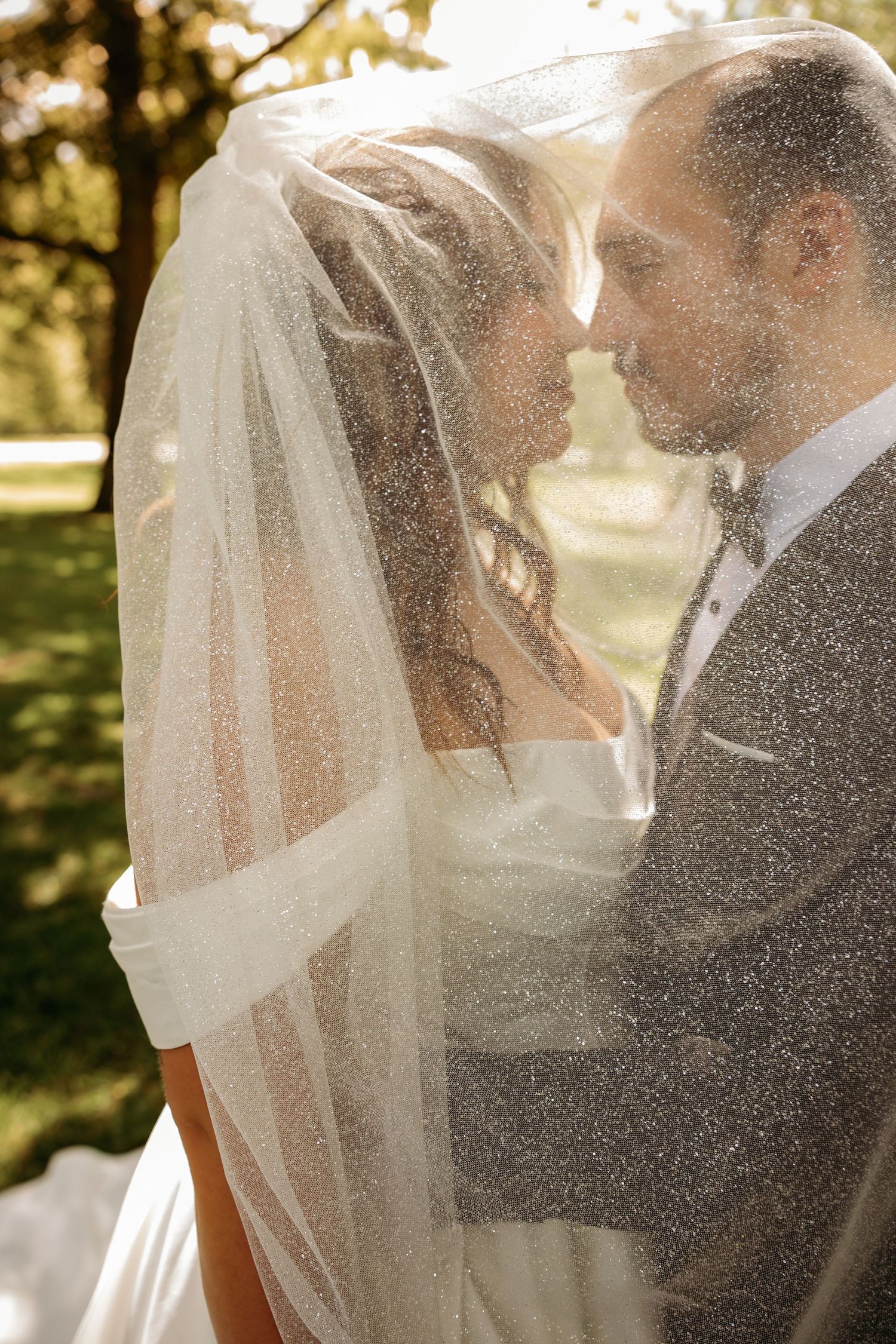Bride and groom sharing a romantic moment under a glittering veil outdoors on their wedding day.