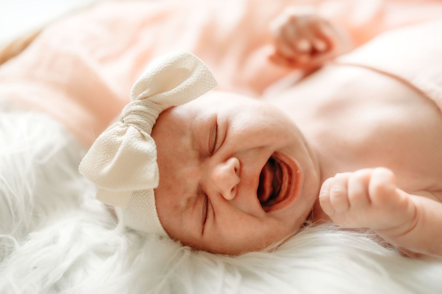 A crying newborn baby with closed eyes lying on white bedding against a pink background.