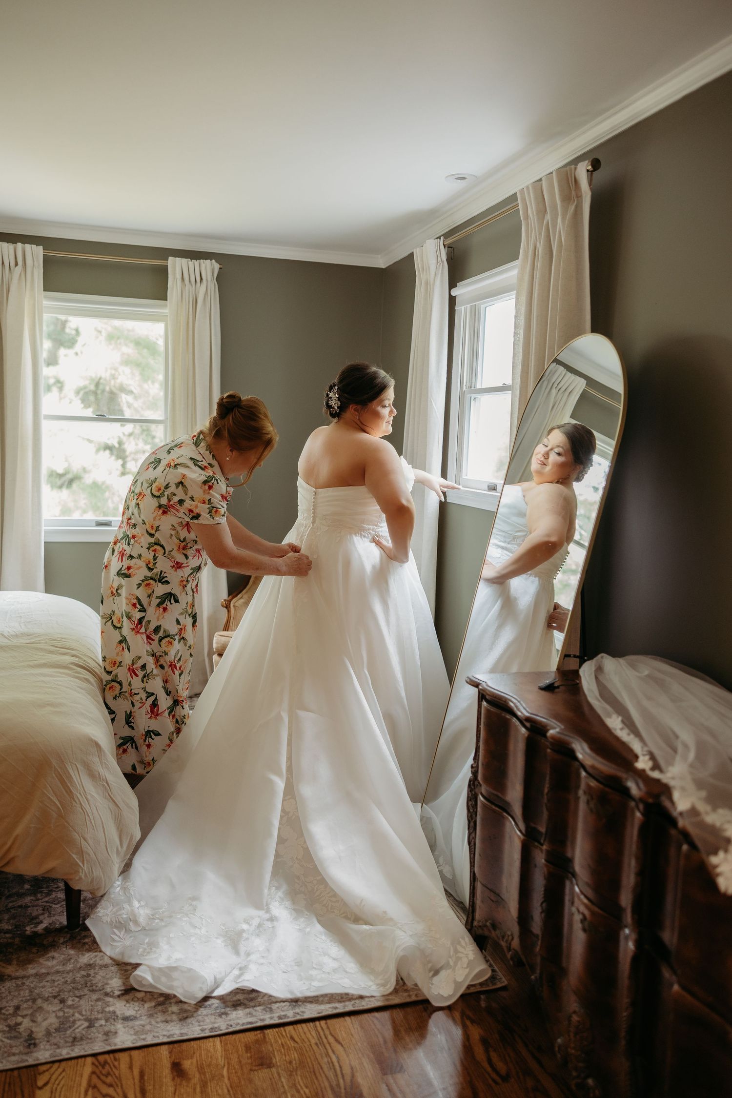 A bride in a white gown gets help from a woman in a floral dress, reflected in a mirror in a cozy bedroom.