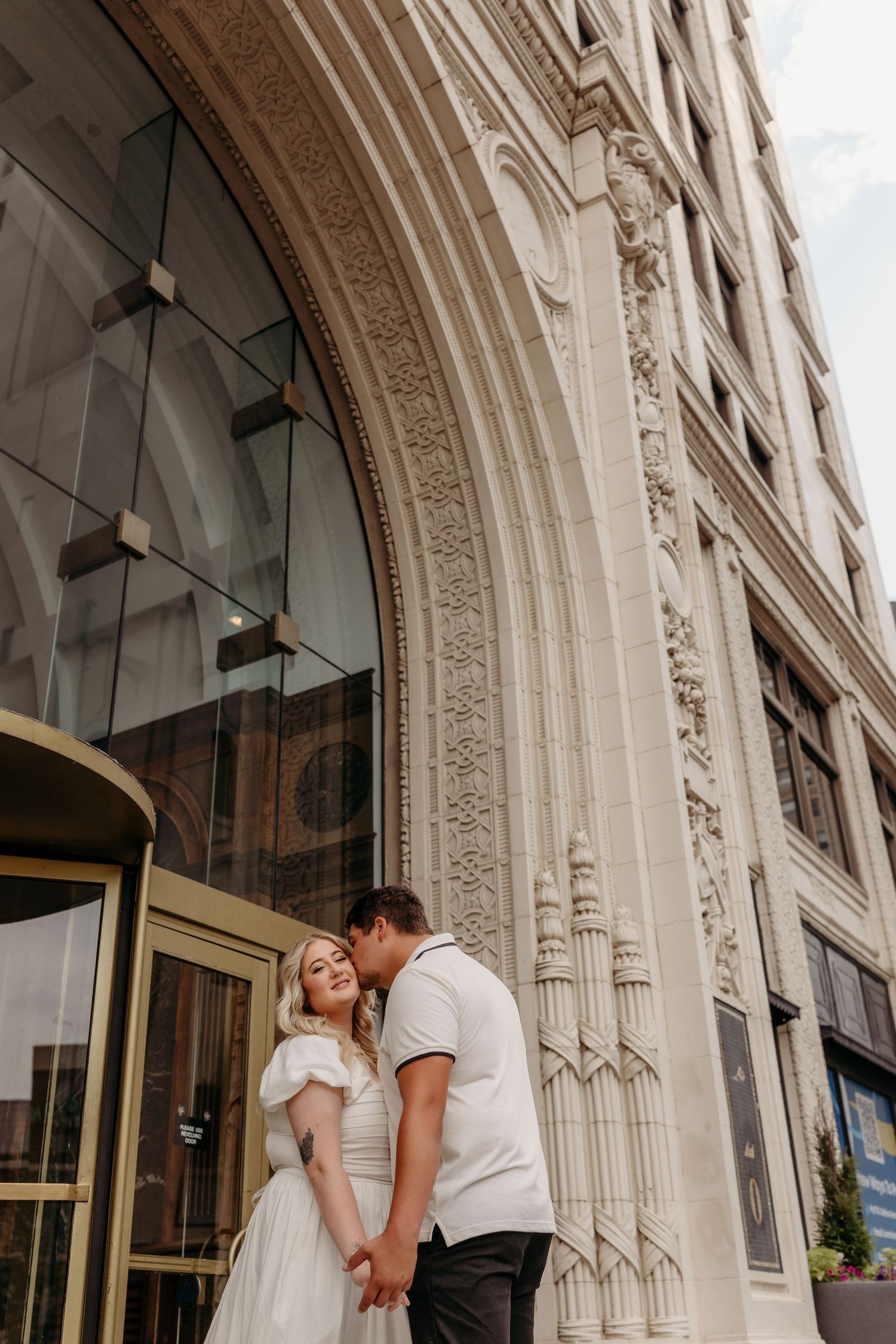 A romantic couple embraces and kisses outside an ornate stone building with Gothic architectural details.