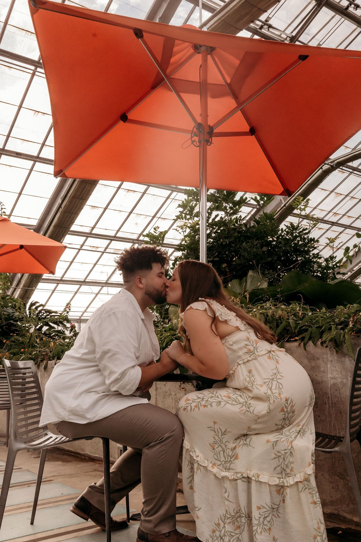 A romantic moment under coral-colored patio umbrellas in a greenhouse setting with lush greenery.