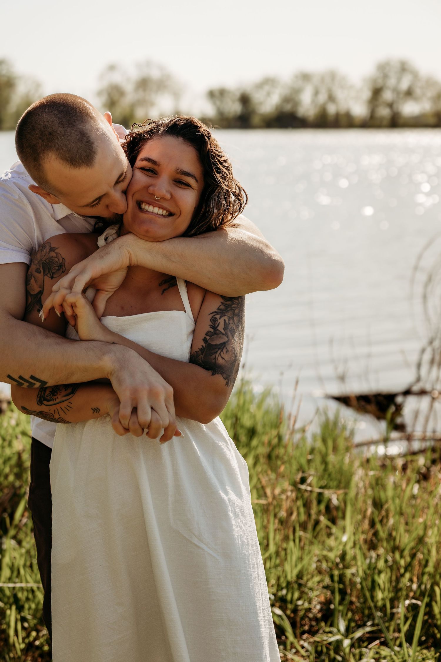 A couple shares an intimate embrace by a lake surrounded by tall grass during a golden hour photoshoot.